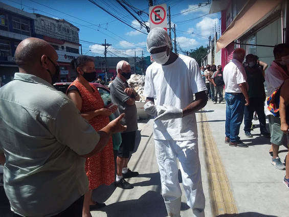 Rapaz vestido com roupa e touca brancas fala com  as pessoas que estão na fila